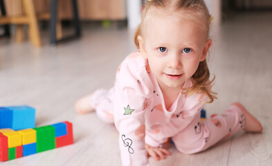 A laughing little child, a blonde preschooler playing with colorful cubes, sitting on the floor in pink pajamas, in a sunny room with a large window at home. Early development of children.