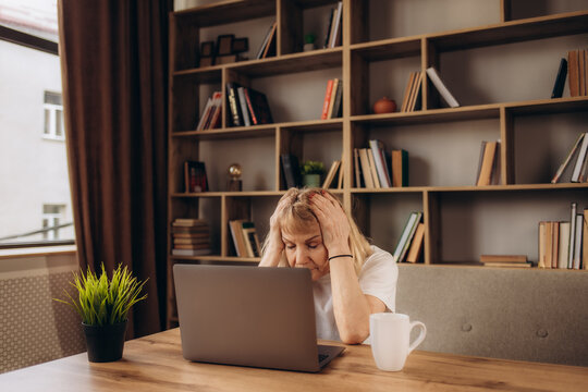 Tired Senior Woman Sitting At Table And Doing Self Head Massage. Exhausted Mature Lady Who Has Headache After Working On Computer For A Long Time Sitting With Her Eyes Closed And Rubbing Her Temples