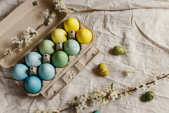 Stylish easter eggs and blooming cherry branch on rustic table. Happy Easter! Rustic easter flat lay. Natural painted eggs in paper tray and spring blossom on linen fabric