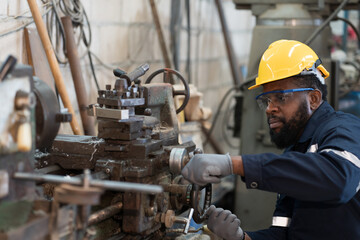 Fototapeta na wymiar Male factory worker working at work maintenance machine in industrial factory while wearing safety uniform, glasses and hard hat. Black male technician and heavy steel lathe machine in workshop