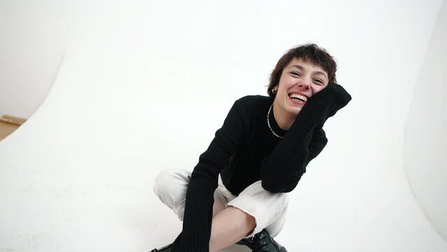 Fashion photoshoot concept. Laughing female model in casual black-and-white outfit sitting on the floor over white background. High quality 4k footage