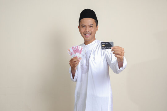 Portrait of attractive Asian muslim man in white shirt with skullcap holding one hundred thousand rupiah and presenting credit card. Isolated image on gray background