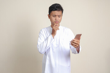 Portrait of attractive Asian muslim man in white shirt thinking about question with hand on chin while holding a mobile phone. Isolated image on gray background