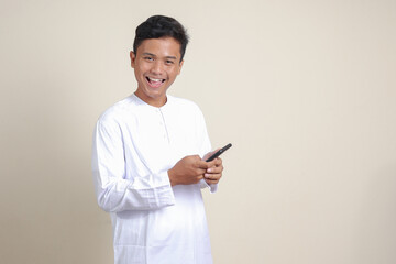 Portrait of attractive Asian muslim man in white shirt holding mobile phone with smiling expression on face. Advertising concept. Isolated image on gray