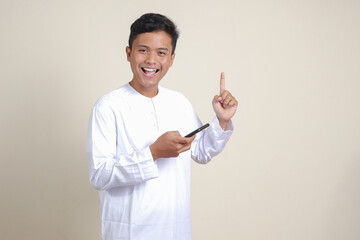 Portrait of attractive Asian muslim man in white shirt holding mobile phone with smiling expression on face while pointing finger to the side. Advertising concept. Isolated image on gray background