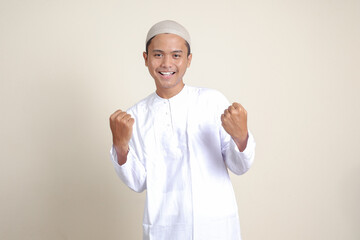 Portrait of attractive Asian muslim man in white shirt raising his fist, celebrating success. Isolated image on gray background