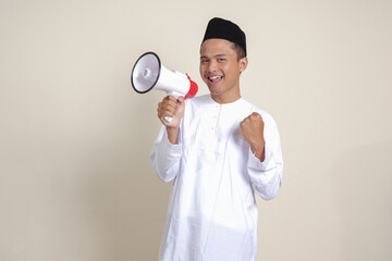 Portrait of attractive Asian muslim man in white shirt with skullcap speaking louder using megaphone, promoting product. Advertising concept. Isolated image on grey background