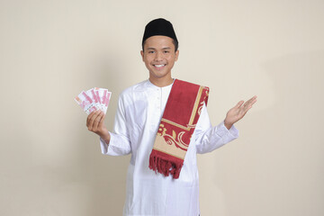 Portrait of attractive Asian muslim man in white shirt showing one hundred thousand rupiah while pointing to the side. Financial and savings concept. Isolated image on gray background