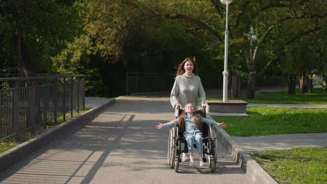 Happy mother pushes daughter in wheelchair running on pavement in city park. Woman and little girl with spinal paralysis enjoy sunny spring day