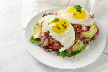 Fried egg, bacon and avocado on a dark whole meal bread with lettuce and tomato, delicious breakfast sandwich, light background, copy space, selected focus