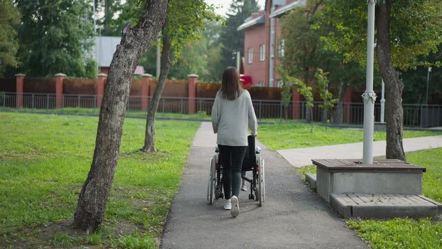 Woman walks on asphalt pavement in park pushing child sitting in wheelchair. Mother and daughter with paralysis enjoy time on fresh air backside view