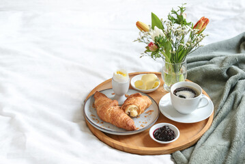 Sunday breakfast in bed with coffee, croissant, jam and boiled egg served on a wooden tray with a bouquet of flowers on mother's day, birthday or valentine's day, copy space