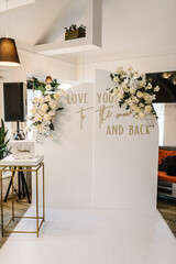 Arch decorated with flowers and greenery with text: love you to the moon and back. Wedding reception for the luxury ceremony. Photo-wall with white box for money or greeting cards in the banquet area.