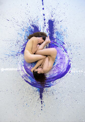 Two nude young sexy women, girlfriends lying like a Yin Yang symbol on the pink, purple, blue and white color painted artists studio floor
