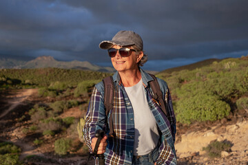 Fototapeta premium Beautiful senior woman in hat and sport clothing in trekking day walking at sunset enjoying freedom and vacation. Happy mature female with stick looking the mountain tropical landscape