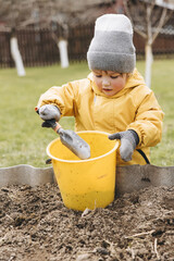 Cute little boy in yellow jacket and bright rubber boots in vegetable garden with big shovel and bucket. Child helping grows plants and vegetables in countryside in spring. Family gardening activity