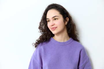 Fototapeta premium Portrait of young beautiful jewish girl with curly dark long hair wearing yellow eye shadow and purple knitted sweater smiling and looking away while standing isolated on white background in studio.