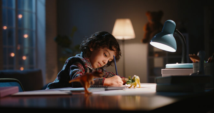 Funny little asian kid drawing at home. Boy with curly hair using his imagination, creating art in evening at home, dreaming of becoming artist - childhood dream, hobby concept 