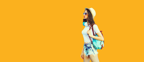 Summer colorful portrait of stylish modern young woman wearing straw hat, backpack and headphones walking on orange background, blank copy space for advertising text