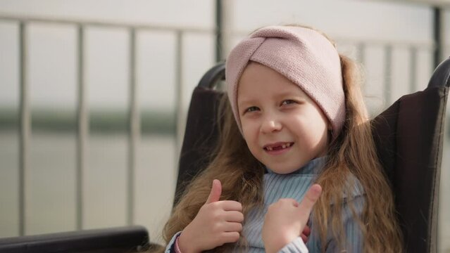 Toothless girl with paralysis turns to camera showing thumbs up gesture on blurred background. Preschooler kid sits in wheelchair on street closeup