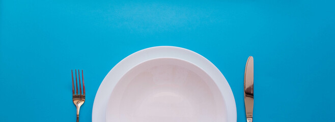  The concept of gastronomy and food. Top view of an empty white plate with fork and knife on a blue...