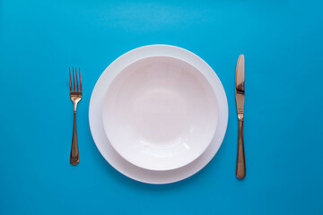  The concept of gastronomy and food. Top view of an empty white plate with fork and knife on a blue background. Serving dinner in a restaurant.