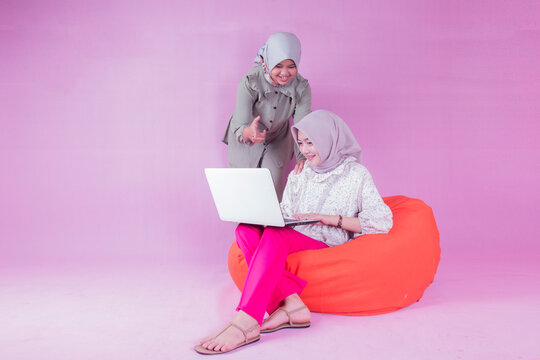 two teenage women in hijab are working sitting on bean chair sofa with laptop isolated in pink indoor studio