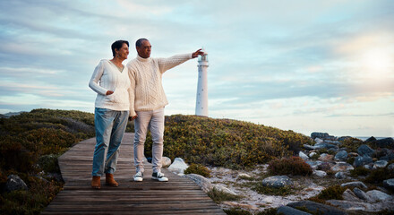 Travel, love and elderly couple pointing on boardwalk at beach, calm at a lighthouse against sunset sky. Senior, man with woman on ocean trip, holiday or vacation, happy and enjoying retirement