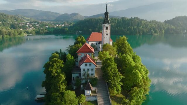 Flying around small island on Bled Lake in Slovenia. Aerial view of Church of the Assumption of Mary in the center of the lake Bled. Warm morning sun and light fog