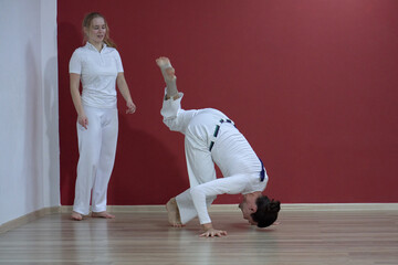 A mature man, a sports master, and a young woman, an aspiring athlete, practice the Brazilian martial art of capoeira in a small gym.