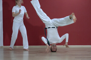A mature man, a sports master, and a young woman, an aspiring athlete, practice the Brazilian martial art of capoeira in a small gym.