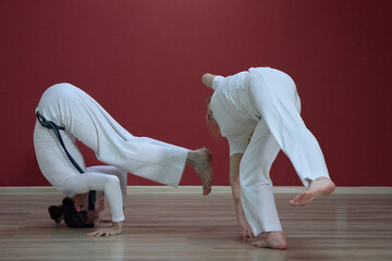 A mature man, a sports master, and a young woman, an aspiring athlete, practice the Brazilian martial art of capoeira in a small gym.