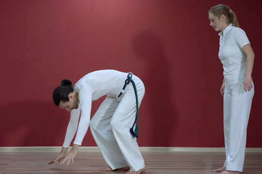 A mature man, a sports master, and a young woman, an aspiring athlete, practice the Brazilian martial art of capoeira in a small gym.
