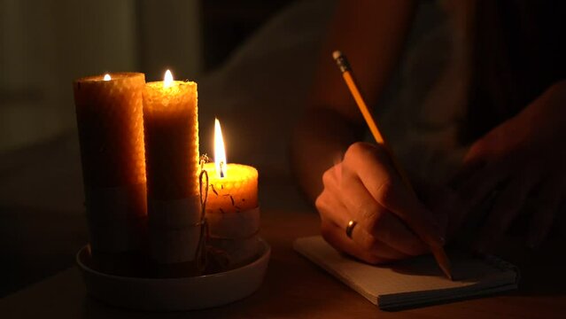 A woman makes notes in a notebook in a dark room with burning candles. Close-up. Magic and occultism.