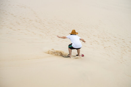 A man riding a sandboard down a dune on the coast of Rio Grande do Sul, Brazil, wearing a straw hat