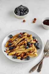 Crepes or blinis or thin pancakes with blueberries, hazelnuts and chocolate sauce on a white plate at white stone table. Maslenitsa traditional Russian festival meal.