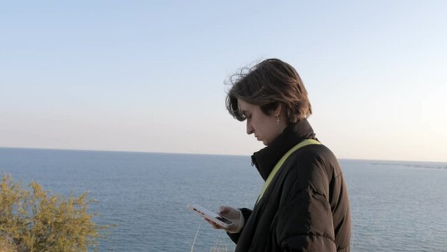 Girl teenager walking on a coastal path and using smartphone, sea ocean in background. Enjoying the time with nature. 