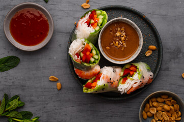 Summer rolls with fresh vegetables and shrimps or prawns and peanut and chili sauce on dark grey background. Vietnamese appetizer. Top view. Healthy food concept