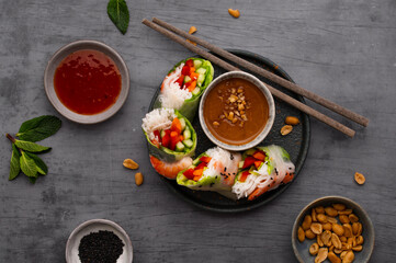 Fototapeta na wymiar Summer rolls with fresh vegetables and shrimps or prawns and peanut and chili sauce on dark grey background. Vietnamese appetizer. Top view. Healthy food concept