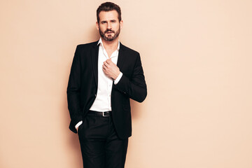 Portrait of handsome confident stylish hipster lambersexual model. Sexy modern man dressed in black elegant suit. Fashion male posing in studio, isolated on beige