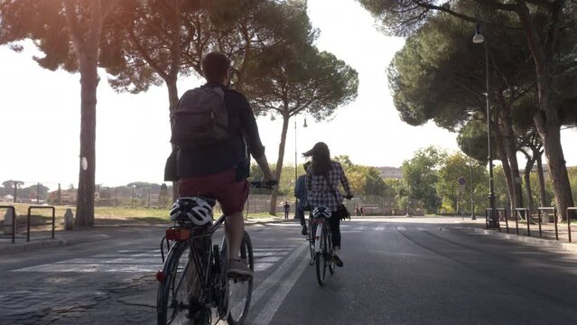 Three young friends tourists riding bikes in colle oppio park on a road with trees in Rome city centre on sunny day slow motion camera car steadycam