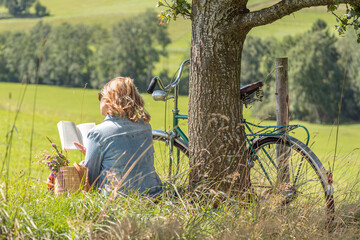 Relaxed blonde Woman reading book under tree. Retro bicycle and basket with flowers.