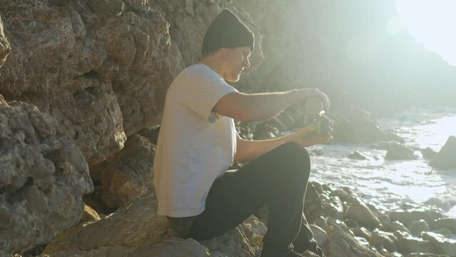 Man unscrews thermos, pours coffee in cup, drinks it, as morning sun shines on him while sea waves crush into rocks near by