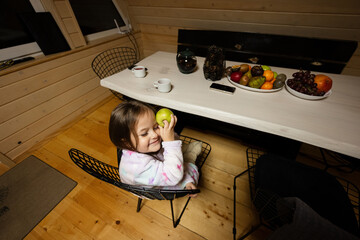 Little cute baby girl in pajama sit kitchen with green apple in cozy wooden tiny cabin house. Life in countryside.