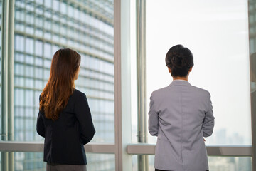 two asian businesswomen standing talking chatting by the window in office