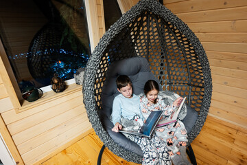 Brother with sister reading books in comfortable hanging chair in cozy wooden tiny cabin house. Life in countryside.
