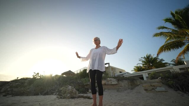 Wide angle view of fit slim mature senior woman stretching practicing yoga and dancing during the morning sunrise on the beach.