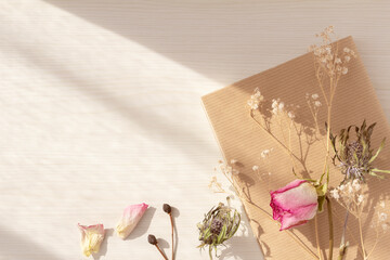 A frame of pink roses, hazel, and dried eryngium flowers accompanying a book on a table at twilight