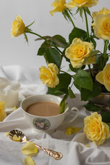 Still-life with porcelain cup of coffee and vintage golden spoon, vase of garden yellow roses, candle on white bed sheet
