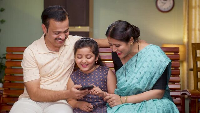 Happy smiling couple with daughter watching mobile phone while sitting on sofa at home - concept of family time, cyberspace and caring parents